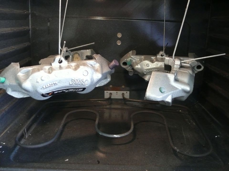 Two silver brake calipers hanging in an oven, one with a black and red sticker that reads 'Brake Performance'