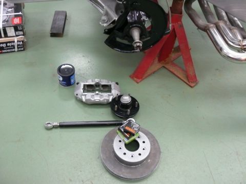 Car brake caliper, rotor, hub, and tie rod on a green floor with boxes and a can