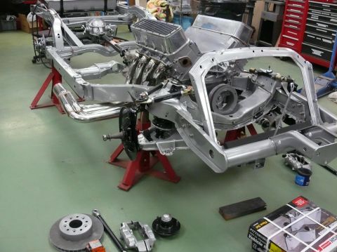 Car chassis on stands with engine and exhaust headers. Parts like brake rotor and caliper are on the floor