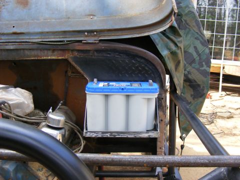 Optima battery mounted in a vehicle's engine bay, with camouflage tarp in background