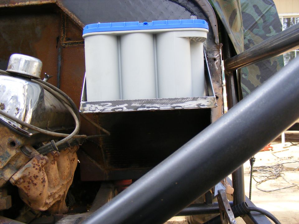 Engine bay with a white battery with blue top mounted on a bracket. Some engine parts and roll cage visible