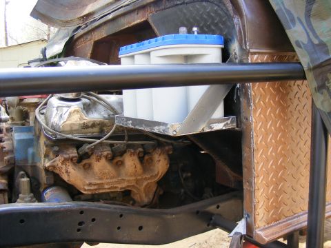 Engine bay with a large white battery in a metal bracket, roll cage, and diamond plate