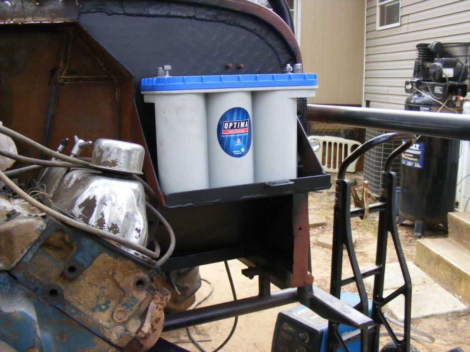 Engine block with chrome valve cover and Optima battery pack mounted on a custom bracket. Jeep grille visible in background