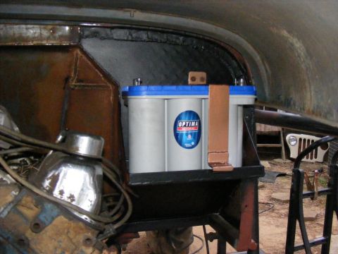 Optima battery mounted in a vehicle, with engine parts visible. Another vehicle's front end is in the backgrou