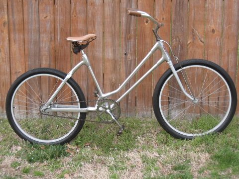 Silver bicycle with tan seat and brown handlebars parked in front of a wooden fence