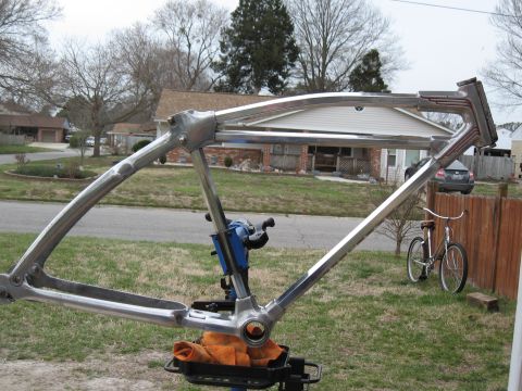 Chrome bicycle frame on a stand, with a complete bicycle leaning against a fence in the background