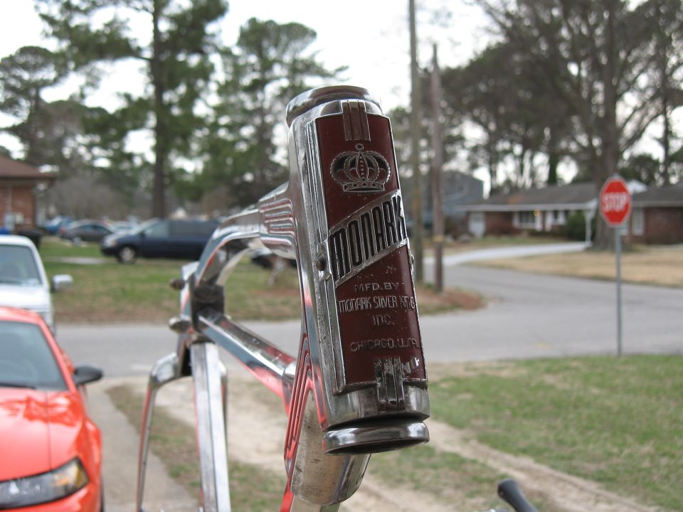 Monark bicycle head badge with crown emblem and text, chrome handlebars, red car in foreground, houses and trees in background