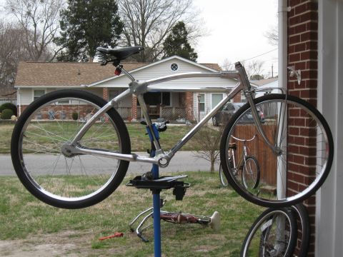 Chrome bicycle frame mounted on a blue repair stand, with another bicycle visible in the background reflection