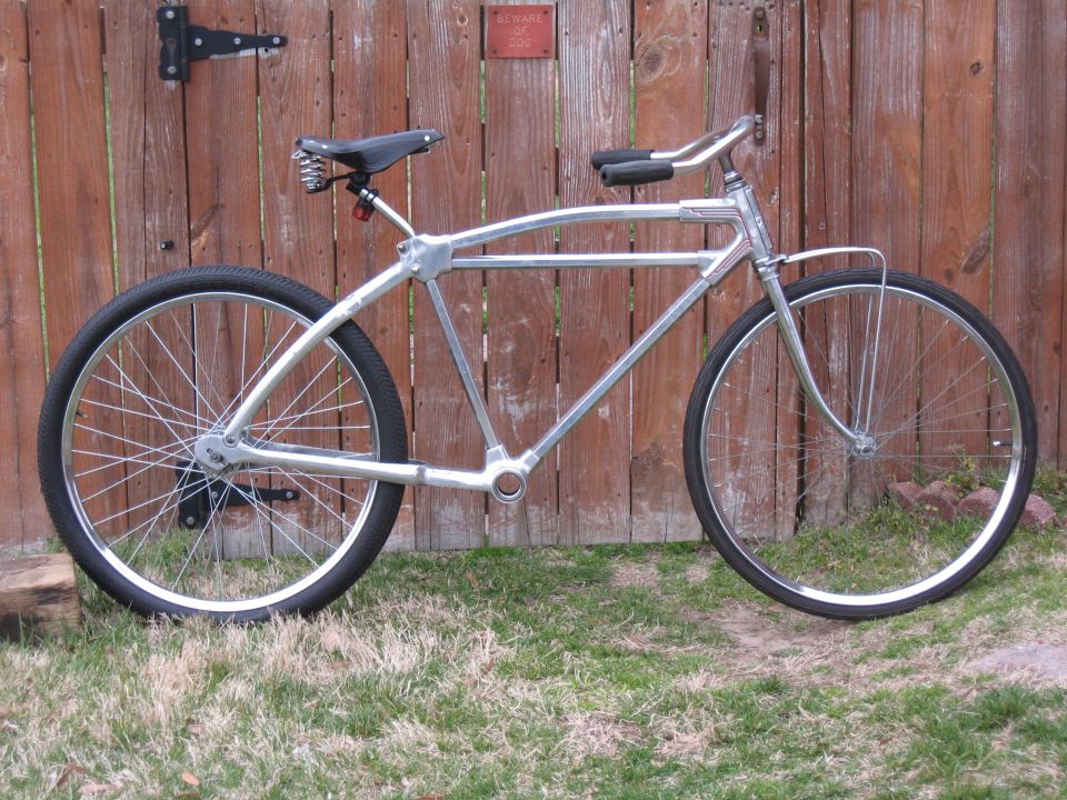 Custom silver cruiser bicycle with wide tires and a spring seat, parked in front of a wooden fence