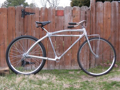 Chrome bicycle frame with black seat and handlebars, parked against a wooden fence