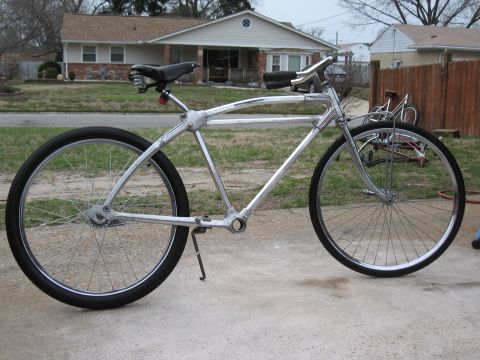 Custom bicycle with chrome frame and black seat, parked on concrete in front of a house. Another bicycle is vi