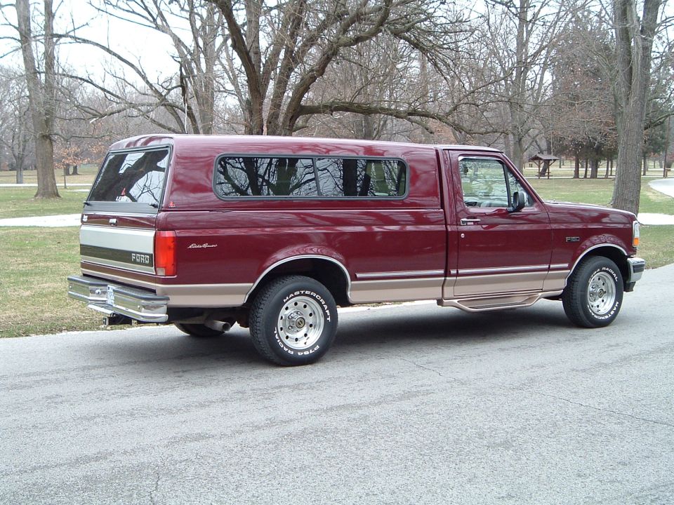 Maroon Ford F-150 pickup with tan two-tone and matching camper shell, rear three-quarter view on a road by a park