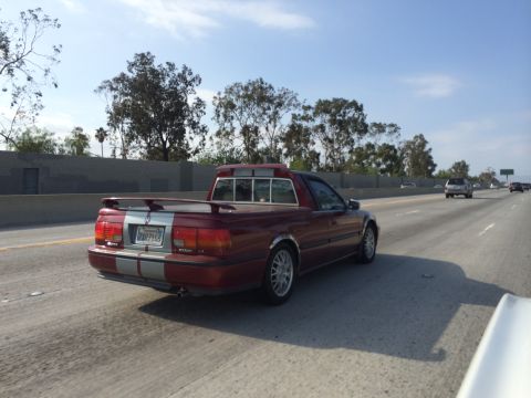 1968 Ford Mustang - A red pickup truck with racing stripes is seen from behind on a sunny day on the highway.
