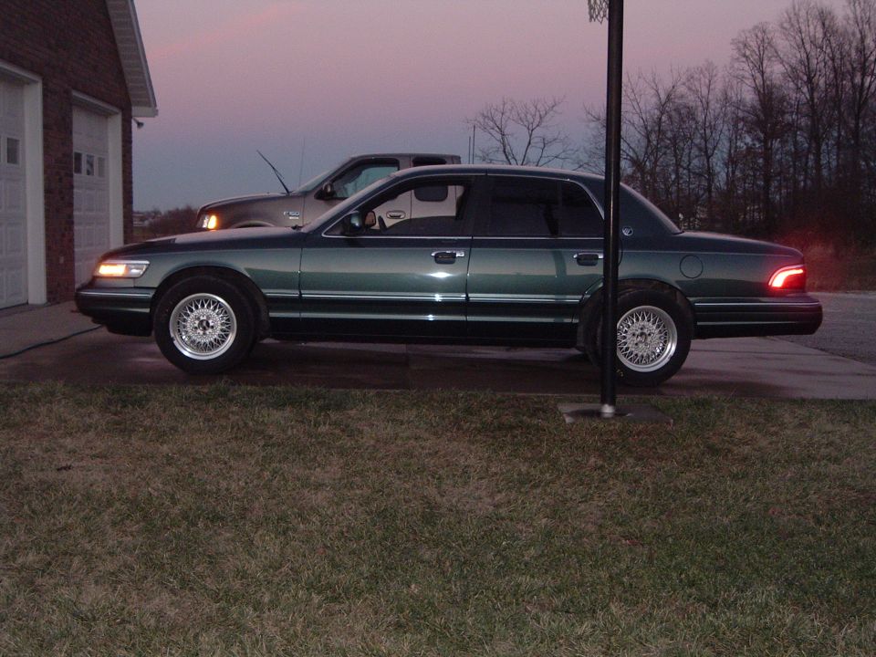 Dark green Mercury Grand Marquis sedan side view at dusk with lights on, parked on driveway