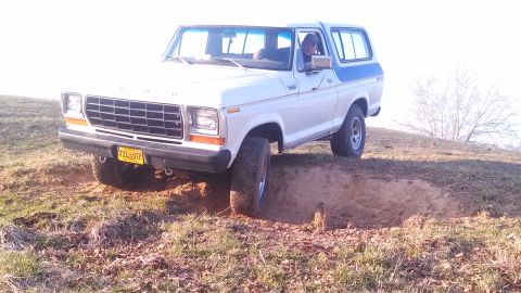 1979 Ford Bronco - A white Ford Bronco with a blue stripe navigates a dirt track, with one wheel dropping into a rut.