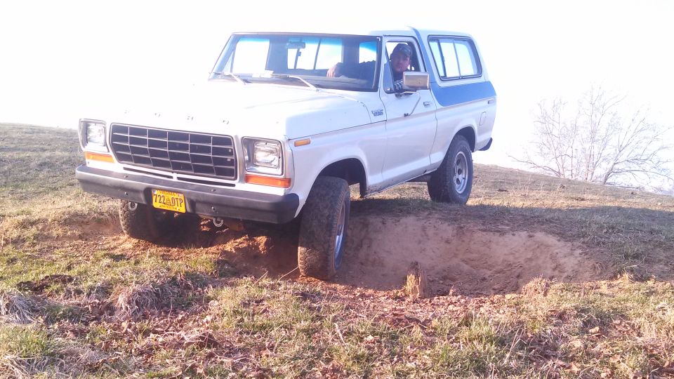 White Ford Bronco with blue trim driving off-road, front driver's side tire in a hole
