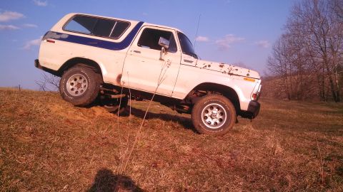1979 Ford Bronco - A white 1978 Ford Bronco with a blue stripe is shown driving on a grassy incline under a clear blue sky.