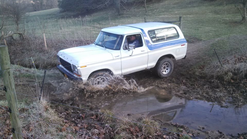 White Ford Bronco with blue stripe driving through a muddy puddle. Driver visible
