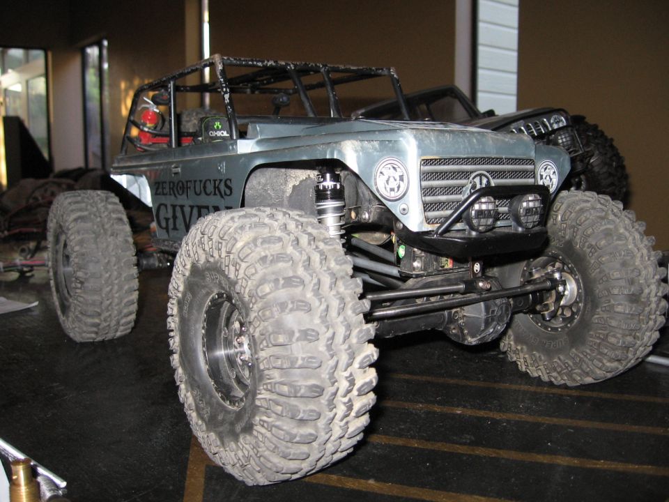 RC rock crawler with large tires and roll cage. Text on the side says 'ZEROFUCKS GIVEN'. Another vehicle is behind it