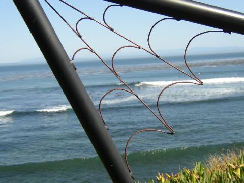 1989 Ford Bronco - Surfers ride waves on a sunny day, viewed through a patterned metal railing overlooking the ocean.