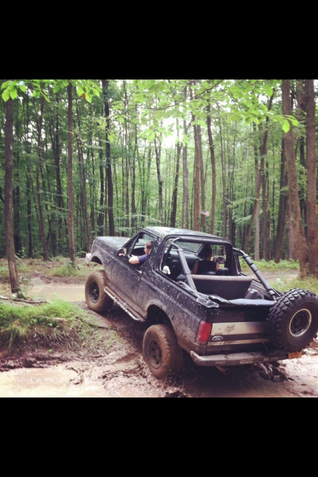 Ford Bronco driving through a muddy trail in a forest. It has a spare tire mounted on the back
