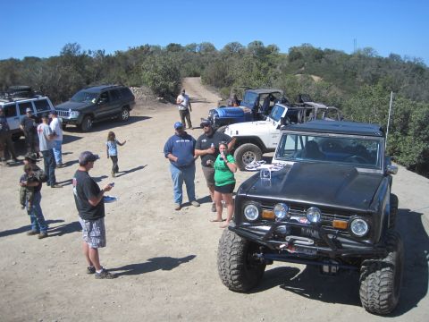 1989 Ford Bronco - A group of people stand around several off-road vehicles on a dirt path.
