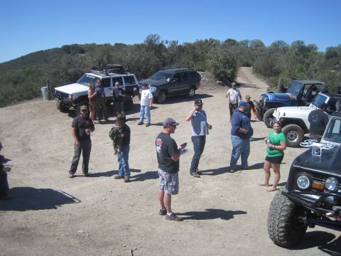 1989 Ford Bronco - A group of people stand around several off-road vehicles in a dirt lot on a sunny day.