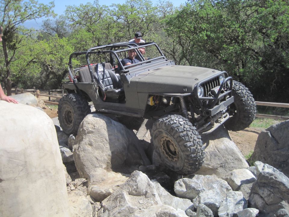 Modified Jeep Wrangler navigating large rocks on a trail. The vehicle has a roll cage and large off-road tires