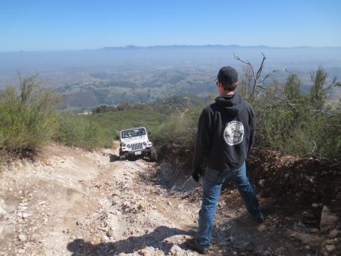 1989 Ford Bronco - A white Jeep Wrangler navigates a challenging off-road trail with a scenic mountain vista in the background.