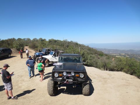 1989 Ford Bronco - A group of people stand around several off-road vehicles parked on a dirt path overlooking a scenic valley.