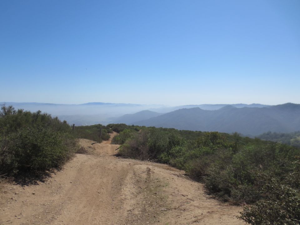 Dirt road winds through dry brush towards hazy mountains under a clear blue sky