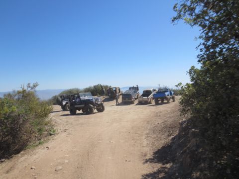 1989 Ford Bronco - A group of off-road vehicles are gathered on a dusty trail under a bright blue sky.