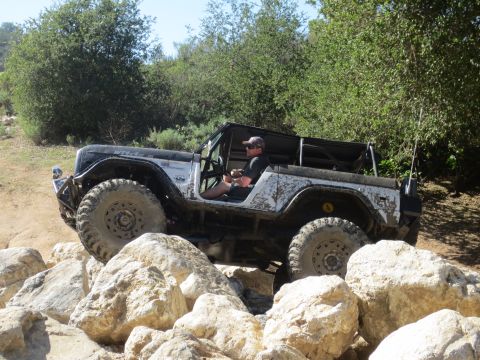 1989 Ford Bronco - A driver maneuvers a modified off-road vehicle over large rocks on a sunny day.