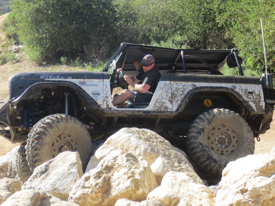 Modified 1978 Ford Bronco with large tires navigating rocky terrain. Driver is visible behind the wheel
