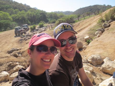 1989 Ford Bronco - Two people pose for a selfie with off-road vehicles navigating a rocky, hilly terrain under a clear sky.