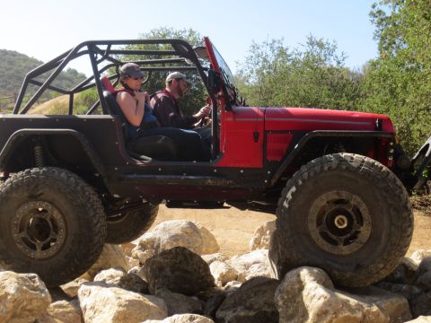 1989 Ford Bronco - A red Jeep Wrangler with a roll cage is shown off-roading over large rocks.
