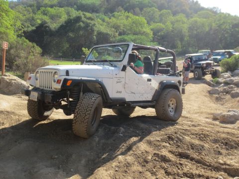 1989 Ford Bronco - A white Jeep Wrangler navigates a rocky, off-road trail with other vehicles in the background.