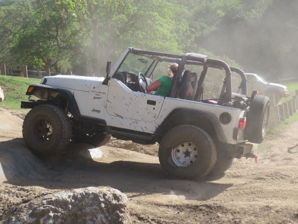 White Jeep Wrangler driving off-road, kicking up dust. Mud splatters on the body