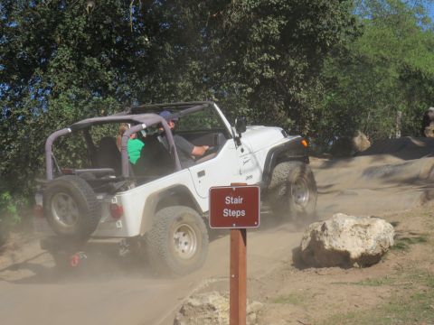 1989 Ford Bronco - A white Jeep Wrangler navigates a dusty off-road trail, passing a sign that reads 'Stair Steps'.