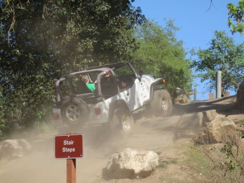 1989 Ford Bronco - White Jeep Wrangler navigates a dusty off-road trail, kicking up dust as it climbs.