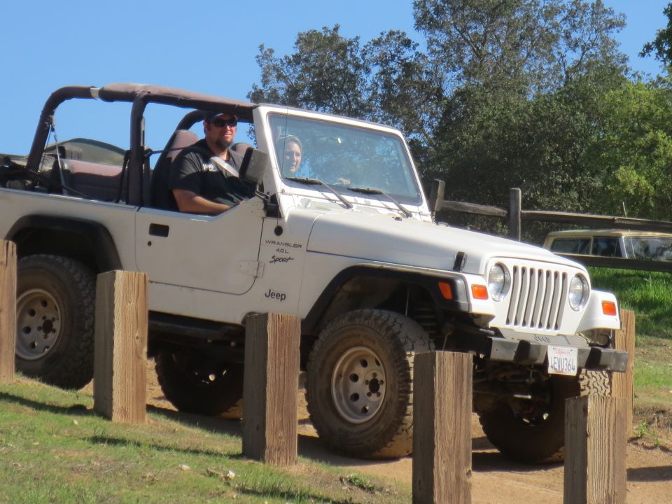 White Jeep Wrangler with soft top removed, driven by a man and passenger, on a dirt path with wooden posts