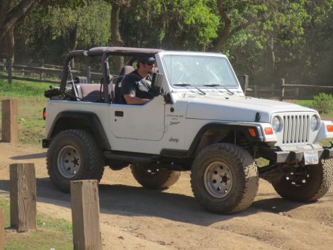 1989 Ford Bronco - A man drives a white Jeep Wrangler on a dirt path surrounded by trees and wooden posts.