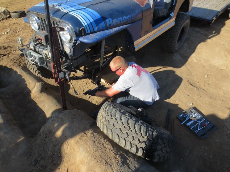 Man working on a blue Jeep CJ-7 Renegade with a Hi-Lift jack and a removed tire. A trailer is attached