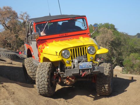 1989 Ford Bronco - A customized Jeep CJ-7 with flame decals navigates an off-road trail under a clear blue sky.