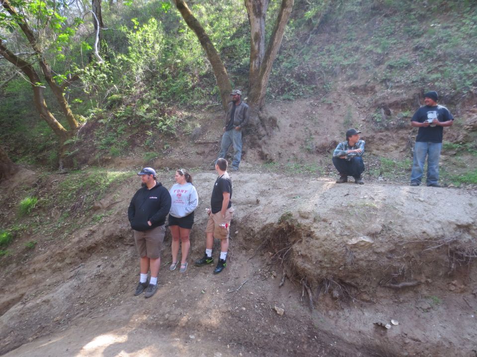 Five people stand and crouch on a dirt embankment with trees and foliage in the background