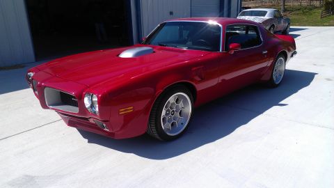 1972 Pontiac Trans Am - A red coupe with a prominent hood scoop is parked in front of a garage, with another vehicle visible behind it.