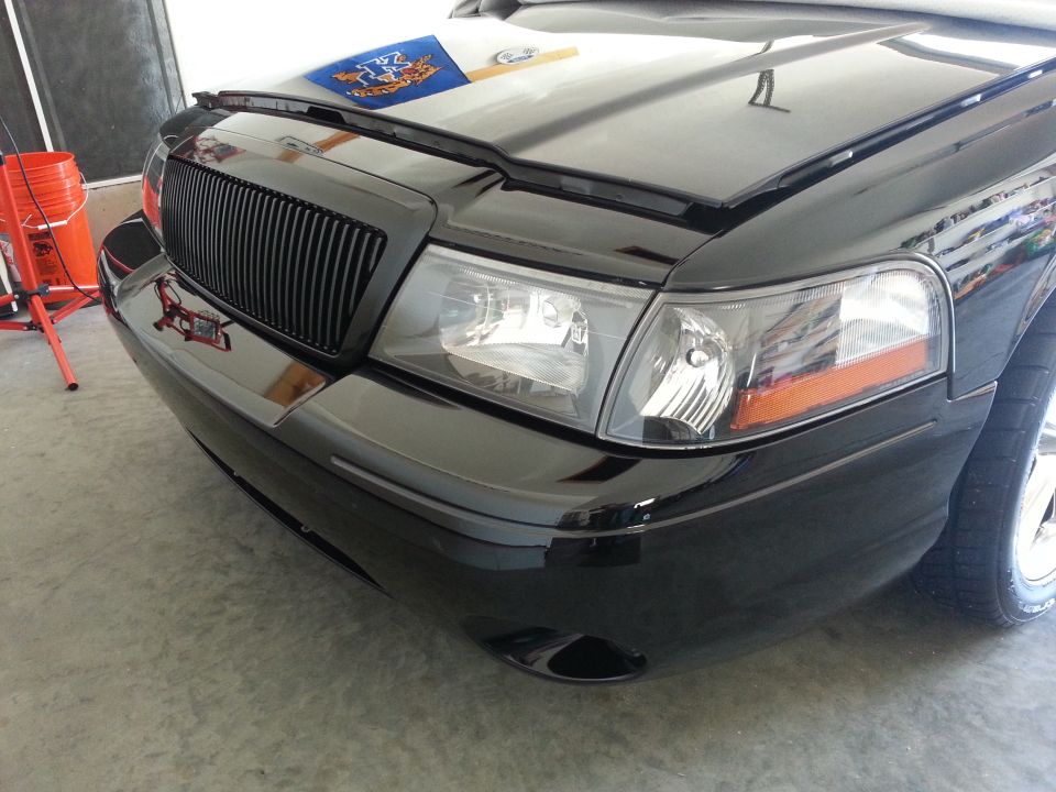 Front of a black sedan with a vertical slat grille and clear headlights. Hood is open, showing a blue and white emblem