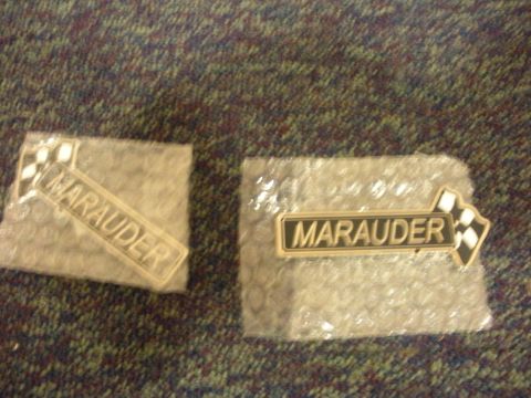 2003 Mercury Marauder - Two Marauder emblems are shown, each with a checkered flag design. The emblems are wrapped in protective plastic.