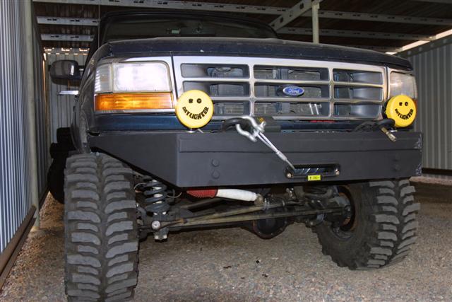 Front of a dark blue 1990s Ford Bronco with winch bumper, smiley KC light covers, solid axle, and big mud tires
