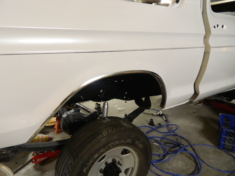 White pickup truck's rear wheel well area, showing suspension and tire. Some blue cords are on the floor
